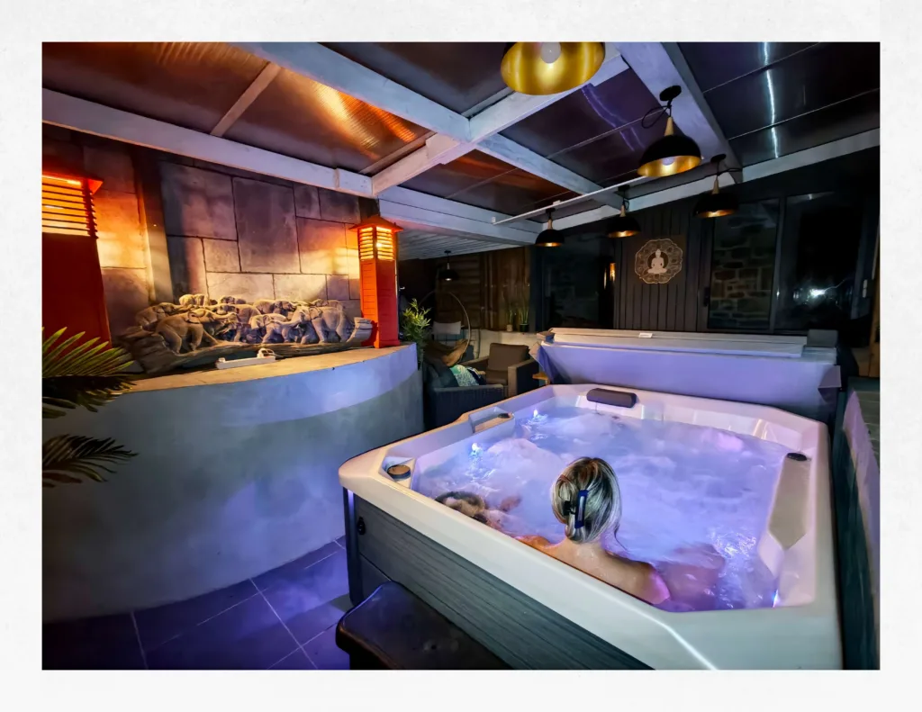 Grand spa jacuzzi privatif 3 places bouillonnant sous le patio couvert et illuminé de la Suite Ananda à Carnac en Bretagne, avec sculpture zen et éclairage romantique.