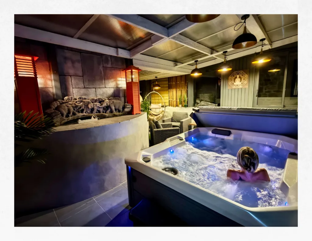 Ambiance nocturne et zen sous le patio couvert de la Suite Ananda aux Suites du Bouddha Bleu, avec jacuzzi 3 places illuminé et salon de détente.
