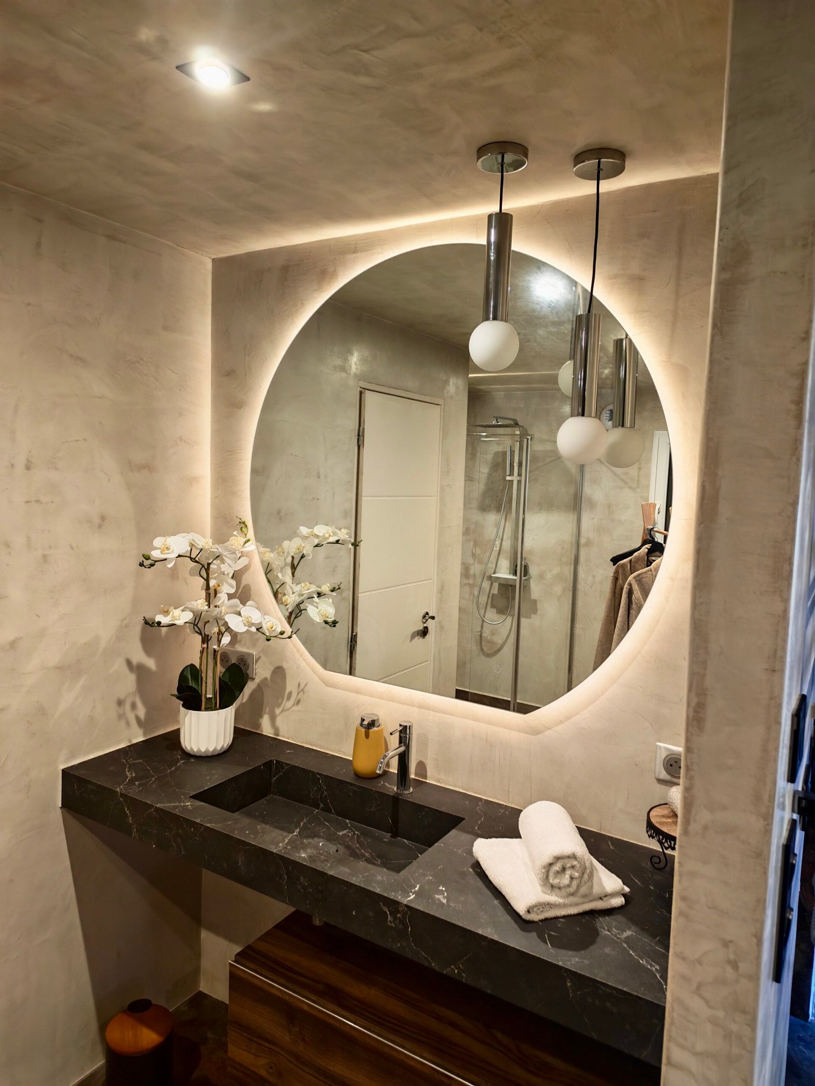 Meuble vasque en pierre noire sur mesure, grand miroir rond rétroéclairé et orchidée blanche dans la salle d'eau de la suite Ananda à Carnac
